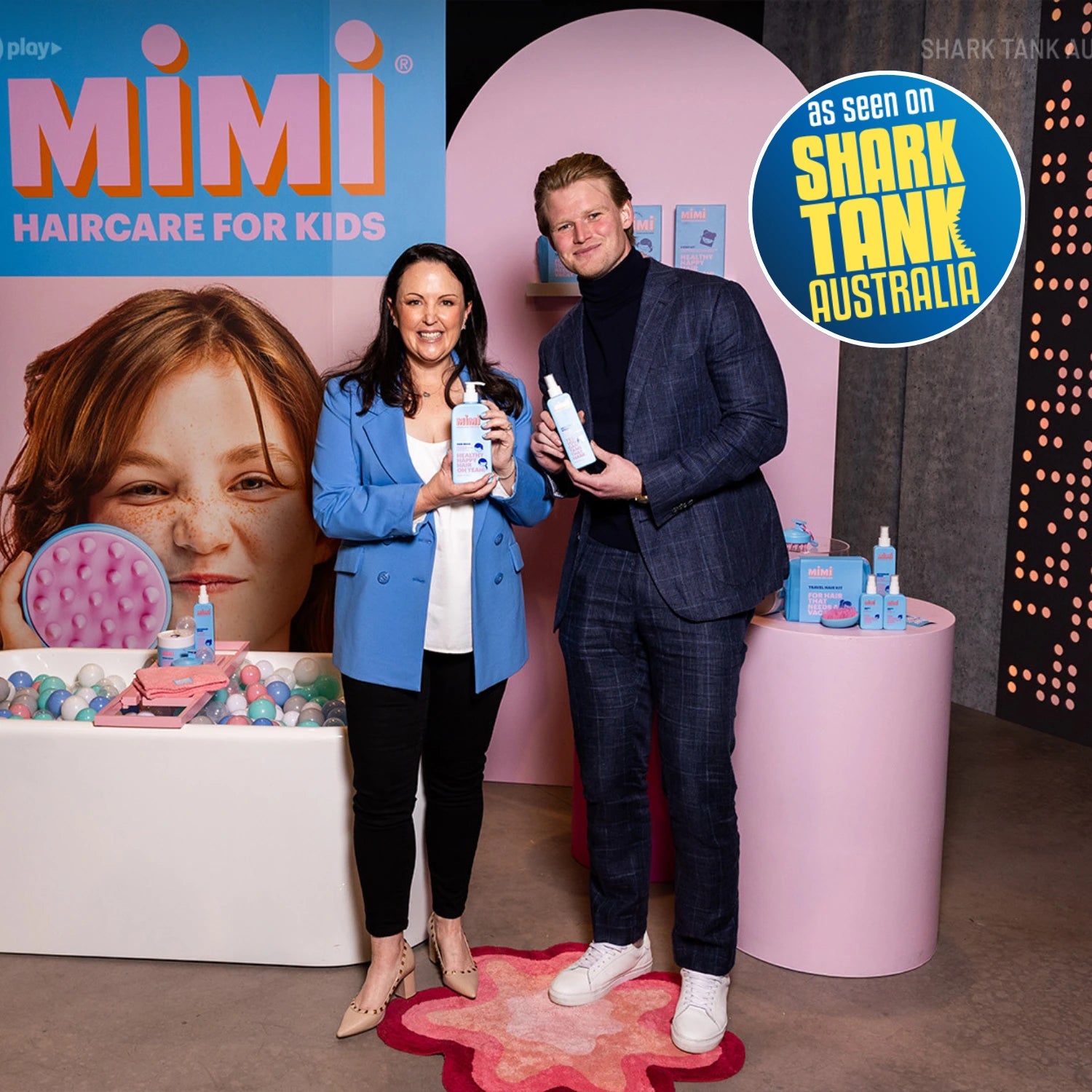 A man and a woman stand in a styled event space holding Mimi Haircare for Kids products. Emma Mackenzie and Davie Fogarty. The background features a pink arch, a large portrait of a child, and promotional graphics including a blue 'Mimi Haircare for Kids' sign and a circular 'As seen on Shark Tank Australia' badge.